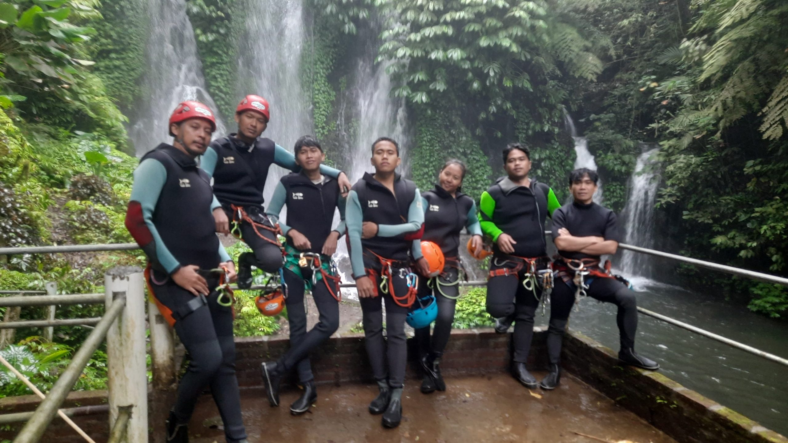 A group of seven canyoning guides wearing wetsuits, harnesses, and helmets pose together on a viewing platform in front of the stunning Last Waterfall at Banyuwana Canyon, surrounded by lush tropical rainforest and cascading waterfalls.