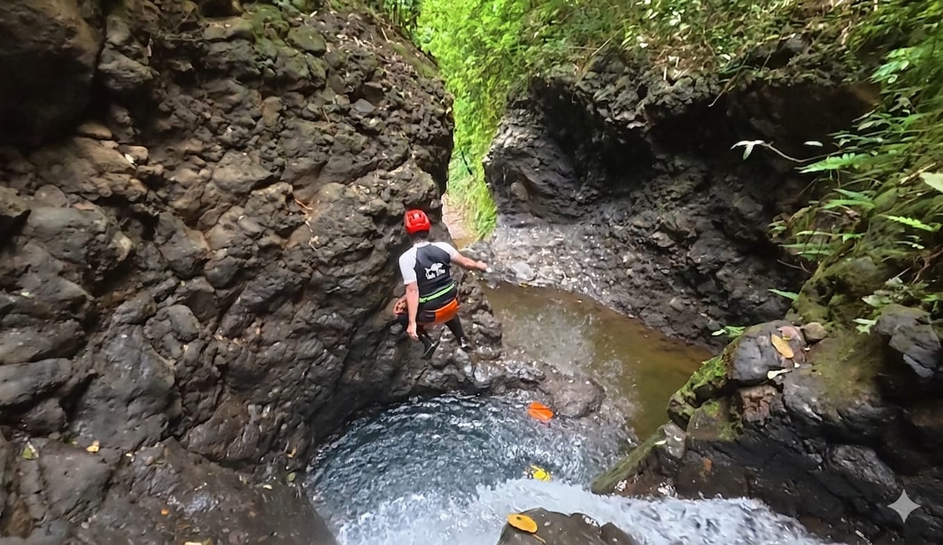 Seorang peserta Canyoning Bali mengenakan helm dan perlengkapan keselamatan sedang melompat ke kolam alami di dalam ngarai yang sempit, dikelilingi tebing batu hitam dan aliran air di lingkungan tropis yang asri.