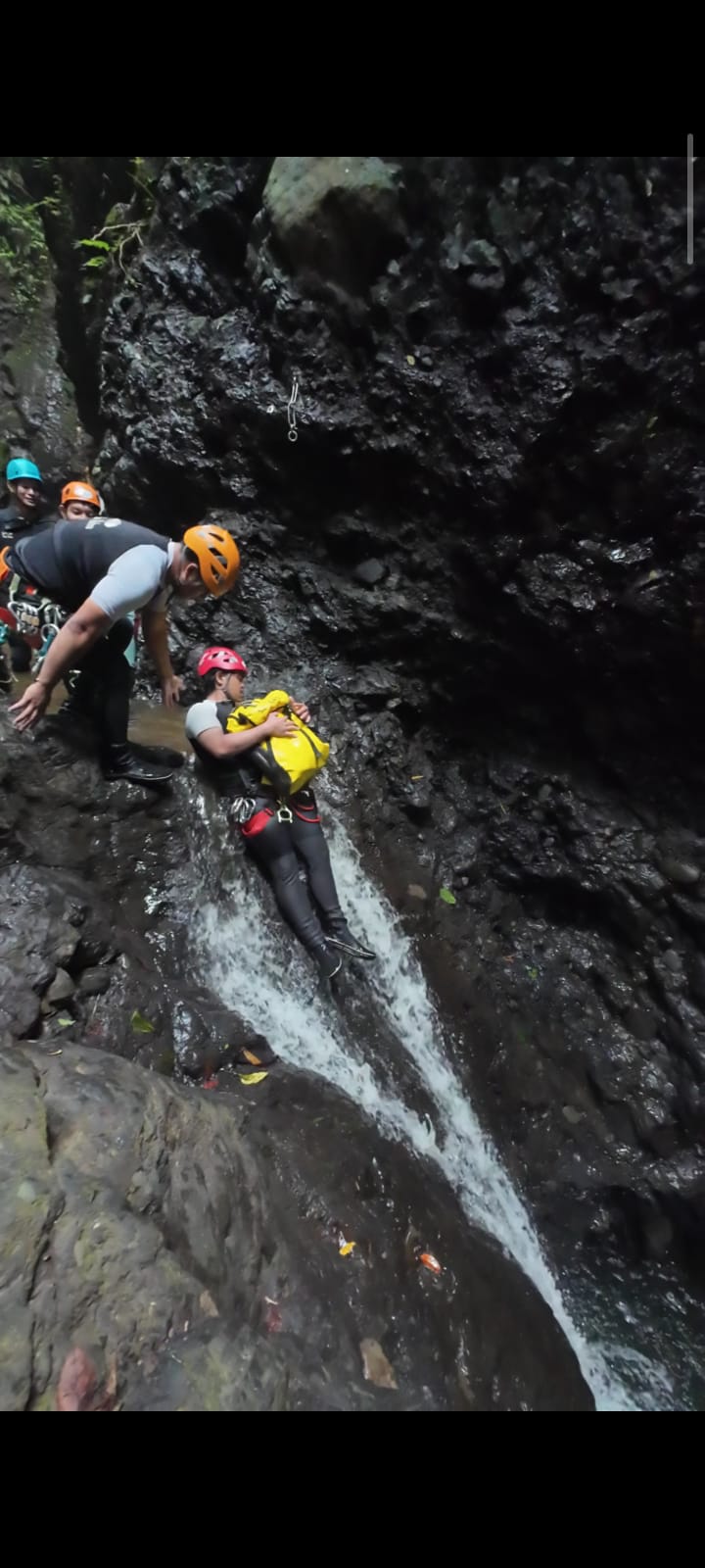 Seorang peserta Canyoning Bali melakukan natural sliding di aliran air sempit di antara tebing batu yang gelap. Peserta mengenakan helm merah, wetsuit hitam, dan perlengkapan keselamatan lengkap, sementara guide bersiaga di samping untuk membantu selama proses sliding alami menuju kolam di bawahnya.