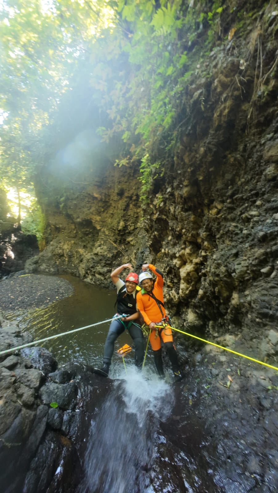 2 orang Canyoning Bali melakukan rappelling di tepi air terjun kecil dengan bantuan guide profesional. Keduanya mengenakan helm, harness, dan perlengkapan keselamatan lengkap sambil berdiri di atas batu vulkanik basah di dalam ngarai yang dikelilingi tebing curam dan vegetasi hijau.