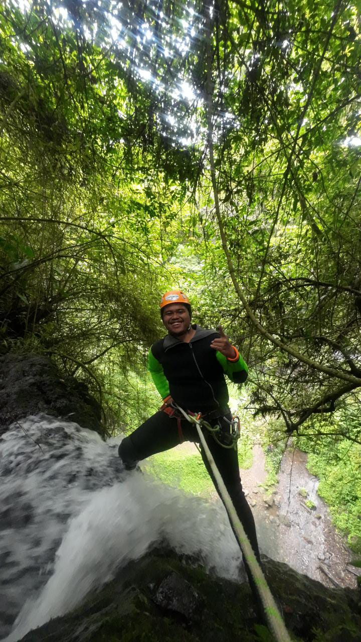 Seorang canyoning guide melakukan rappelling di tepi air terjun dengan menggunakan descender dalam mode cepat. Guide mengenakan helm oranye, wetsuit hitam-hijau, dan perlengkapan keselamatan lengkap sambil mengontrol kecepatan penurunan di atas tebing basah yang dikelilingi vegetasi yang lebat.